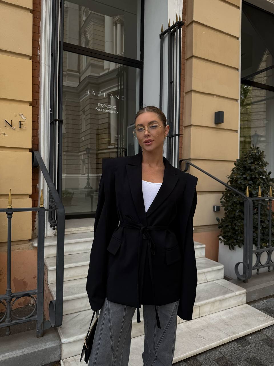 Person wearing a black blazer and gray pants standing in front of a building entrance.