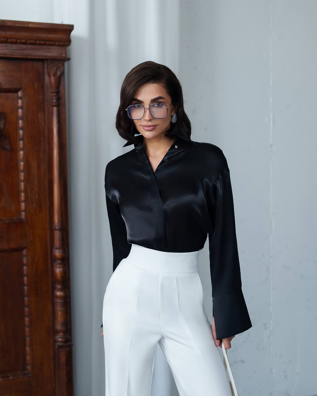 Woman wearing a black satin blouse and white pants against a light gray wall.