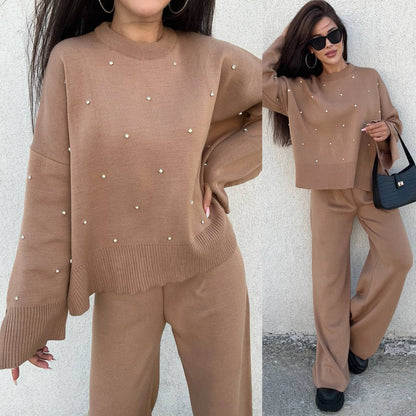Woman wearing a brown sweater and pants set with pearl embellishments, standing against a white wall.