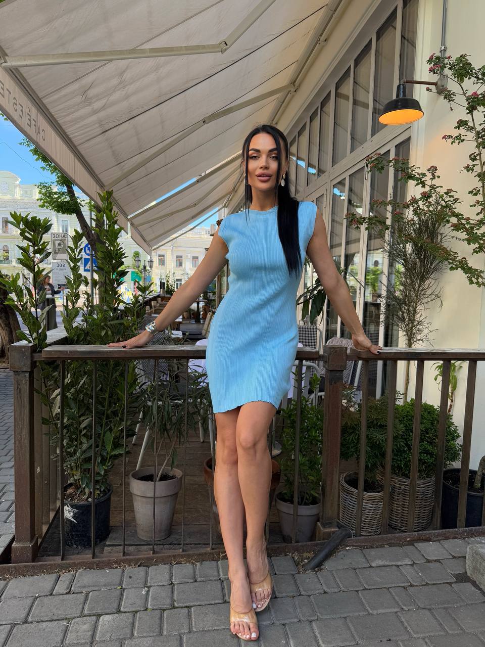 Woman in a light blue dress standing on a balcony with plants and a building in the background
