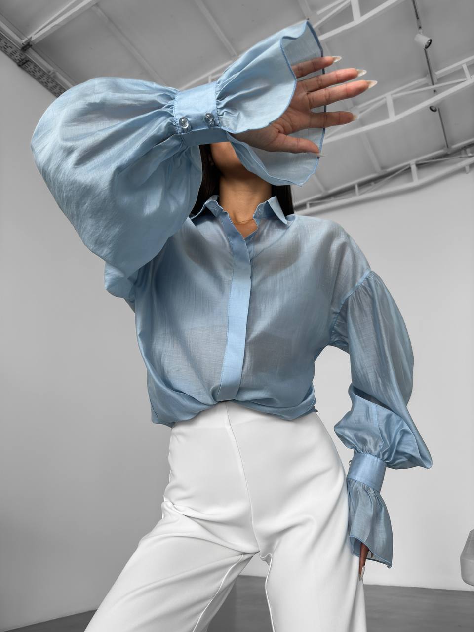 Person wearing a light blue blouse with puffed sleeves and white pants in an indoor setting.