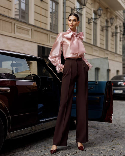 Woman in a pink blouse and brown pants standing next to a black car on a city street.