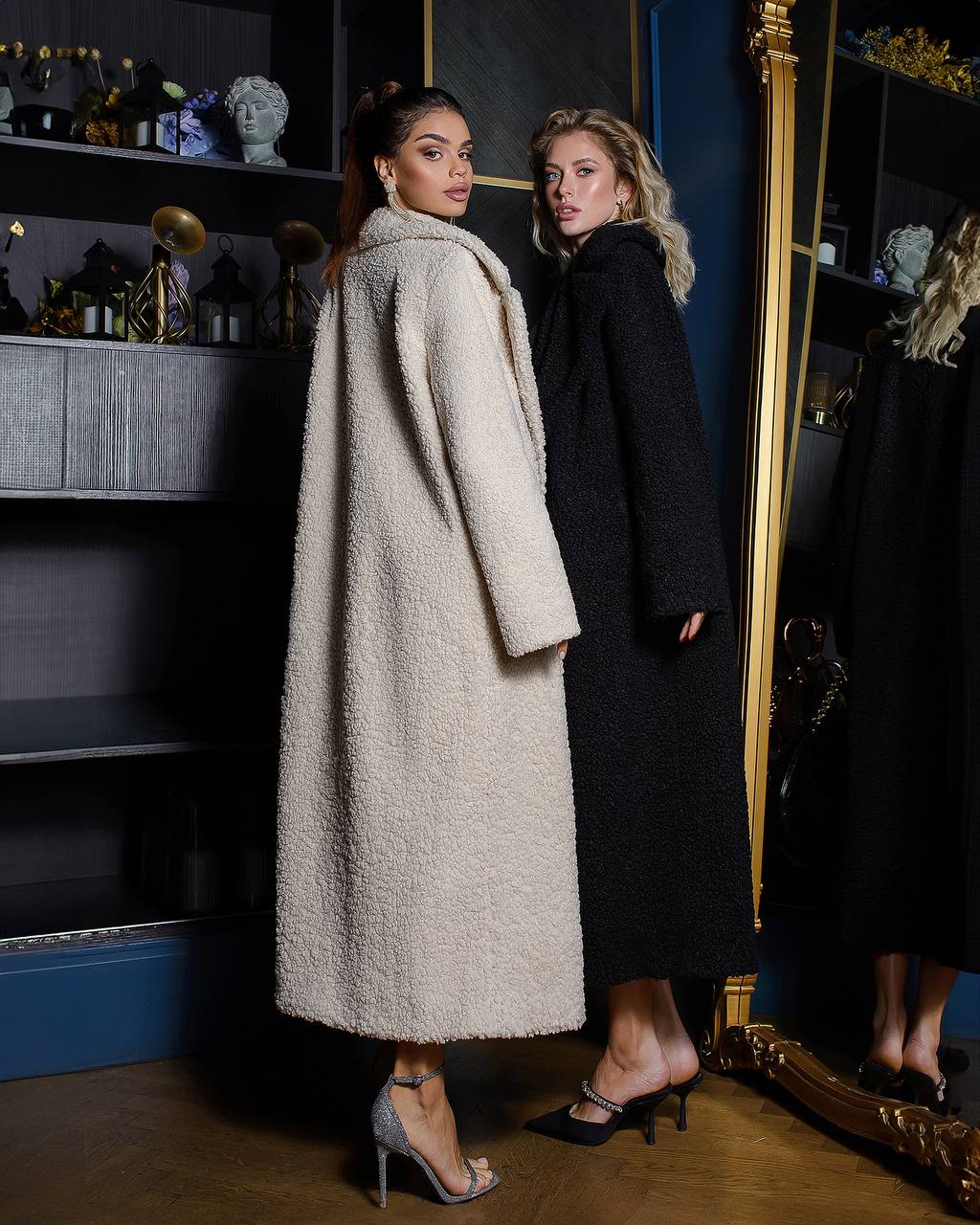 Two women wearing long coats standing in a room with shelves and decorative items.