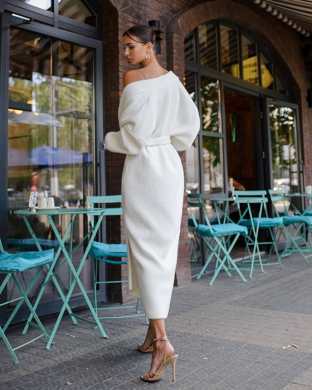 Woman in a white outfit standing outside a building with teal chairs and tables.