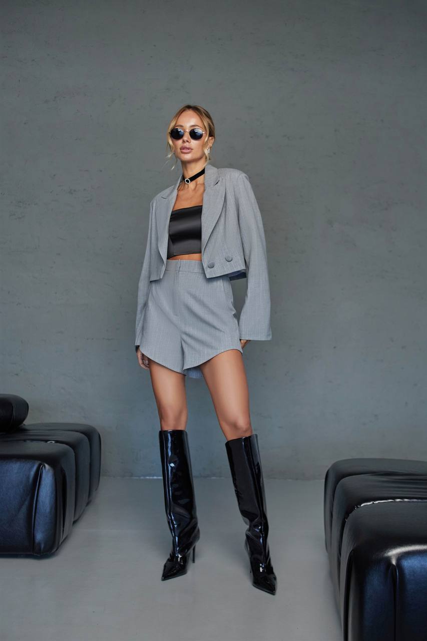 Woman in a gray outfit with black boots standing against a plain background