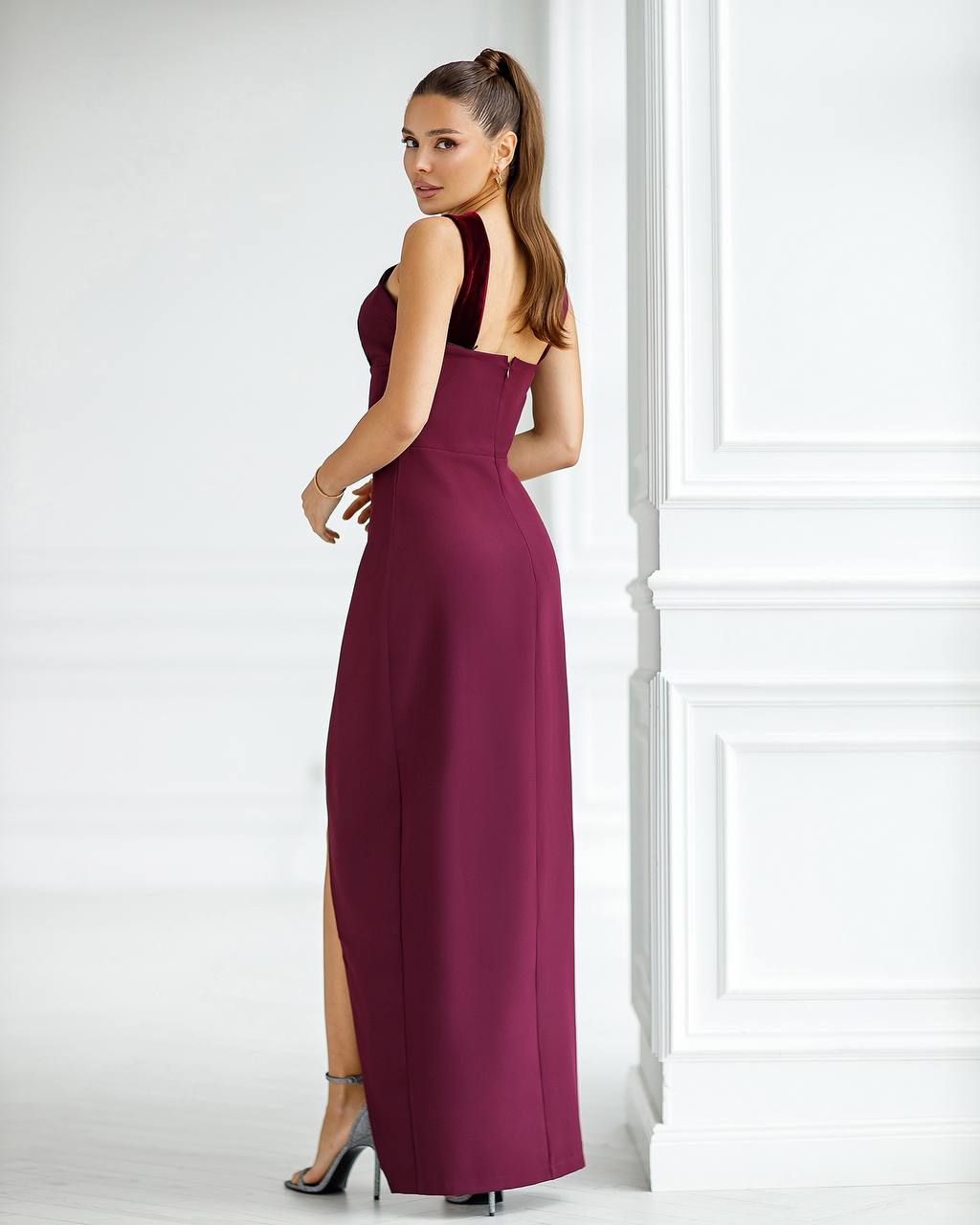 A woman modeling a sleeveless, floor-length burgundy maxi dress with a high side slit, shown from a three-quarter back angle in a bright white room with decorative wall molding.