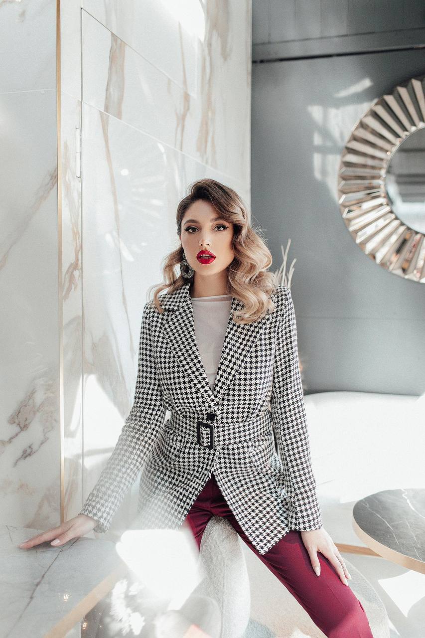 Woman in a checkered blazer and red pants posing in a modern interior setting.