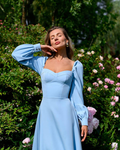 Woman in a light blue dress standing in a garden with flowers and greenery.