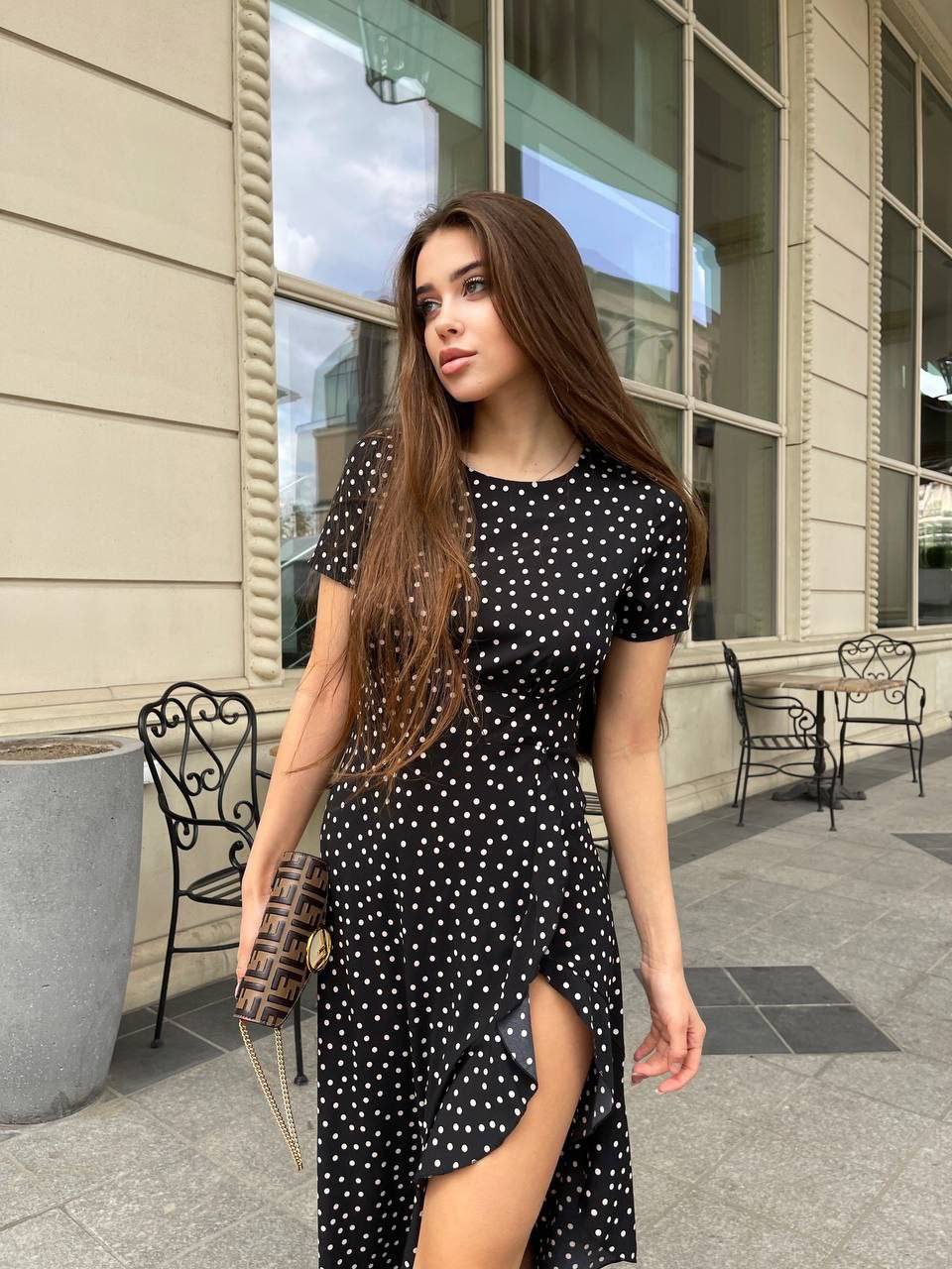 Woman in a black polka dot dress standing outdoors near a building.