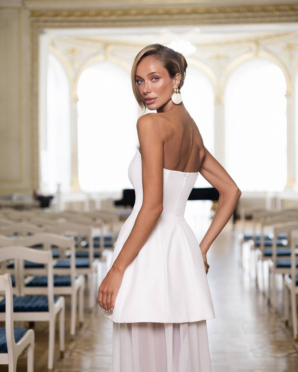 Woman in a white dress standing in an elegant room with chairs and large windows.