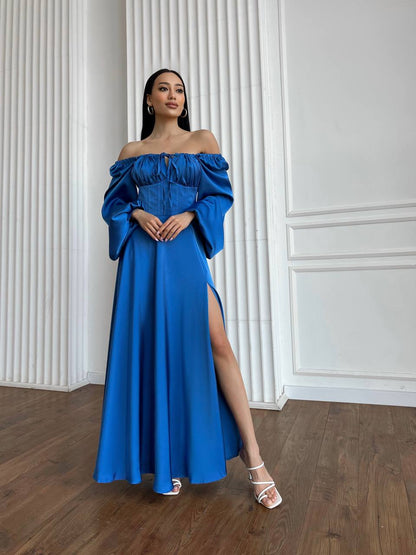 Woman wearing a blue off-shoulder dress with a slit, standing against a white paneled wall.