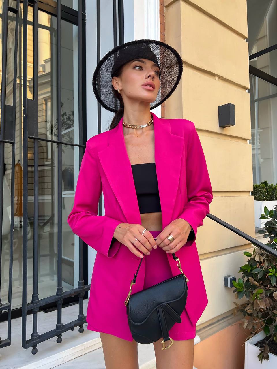 Woman in a bright pink blazer and black hat standing outside a building.
