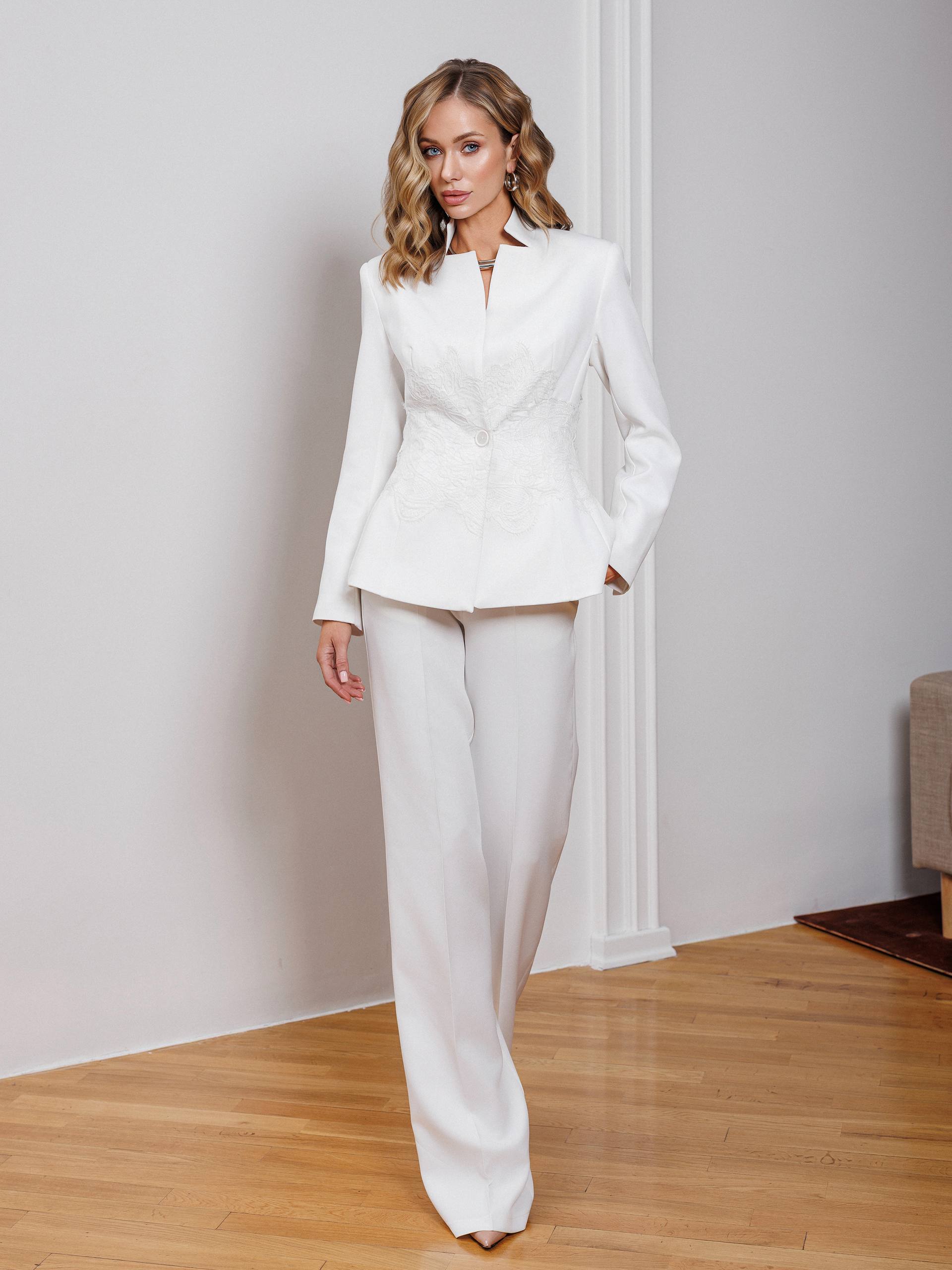 Woman wearing a white suit standing in a room with wooden floor and white walls.