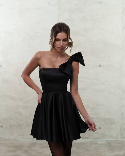 Woman wearing a black one-shoulder dress with a bow detail against a neutral background