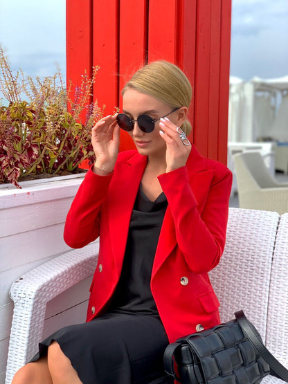 Woman in a red blazer and black dress sitting on a white bench with a red wall and plants in the background.