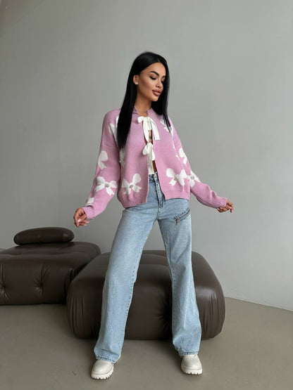 Woman wearing a pink sweater with white floral patterns and blue jeans standing in a minimalistic room.