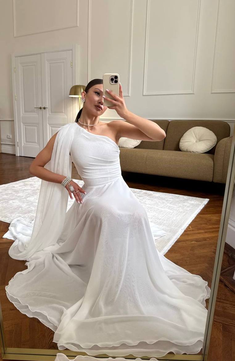 Woman in a white one-shoulder dress taking a mirror selfie in a room with a couch and rug.