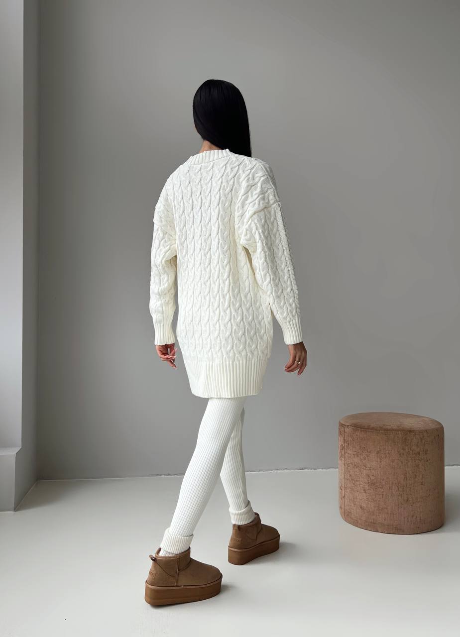 Person wearing a white cable knit sweater and brown boots in a minimalistic setting.