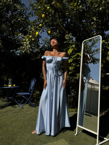 Woman in a light blue dress standing outdoors next to a mirror, with trees and a clear sky in the background.