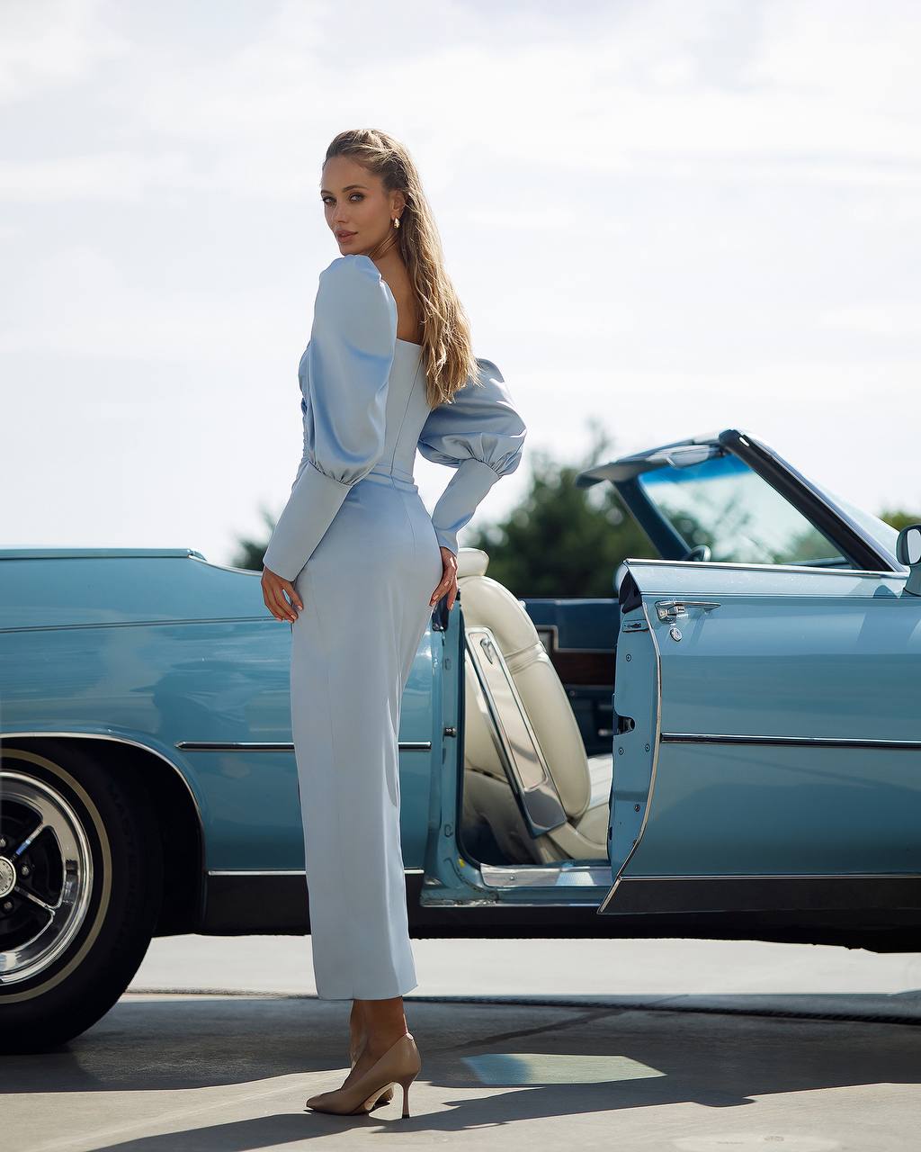 Woman in a white outfit standing next to a vintage blue car with an open door.