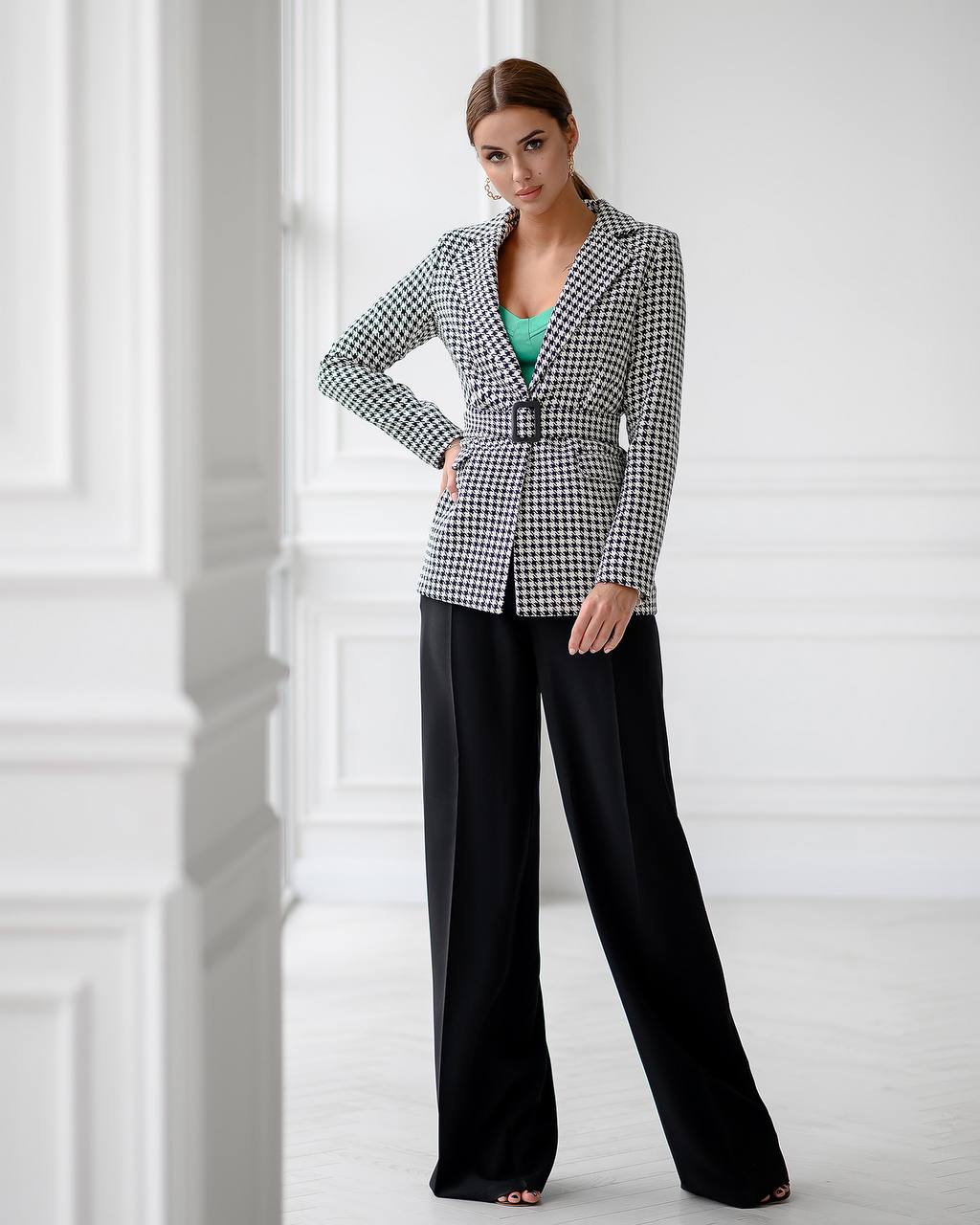 Woman wearing a checkered blazer and black pants in a white room.
