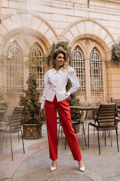 Woman in a white blouse and red pants standing in an outdoor setting with architectural elements.