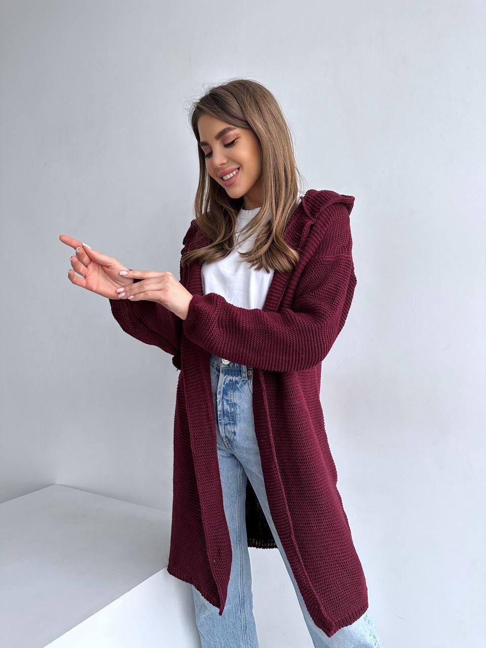 Woman wearing a burgundy cardigan over a white shirt and blue jeans on a plain background