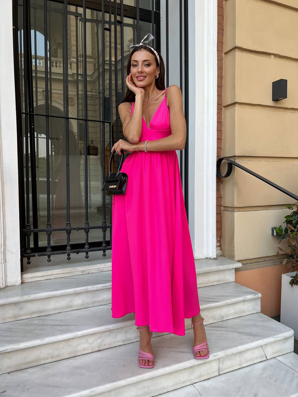 A woman wearing a bright pink sleeveless maxi dress stands on white stone steps in front of a building entrance with a black metal gate and beige wall.