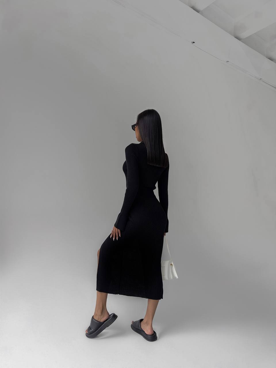 Person wearing a black dress standing in a minimalistic room with white walls and gray floor.