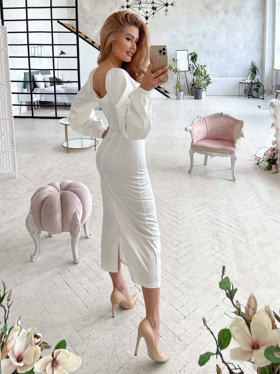 Woman in a white dress taking a selfie in a stylish room with pink furniture and floral decorations.