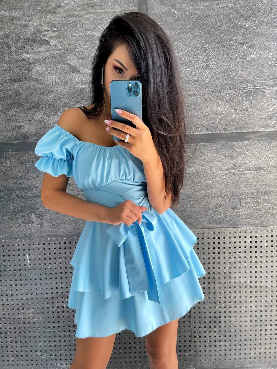 Woman taking a mirror selfie wearing a light blue off-shoulder dress against a gray wall.