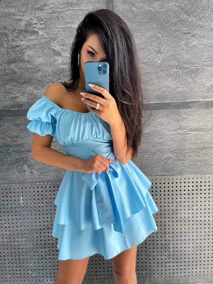 Woman taking a mirror selfie wearing a light blue off-shoulder dress against a gray wall.