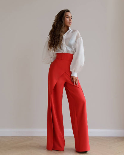 Woman wearing a white blouse and red pants against a plain wall.