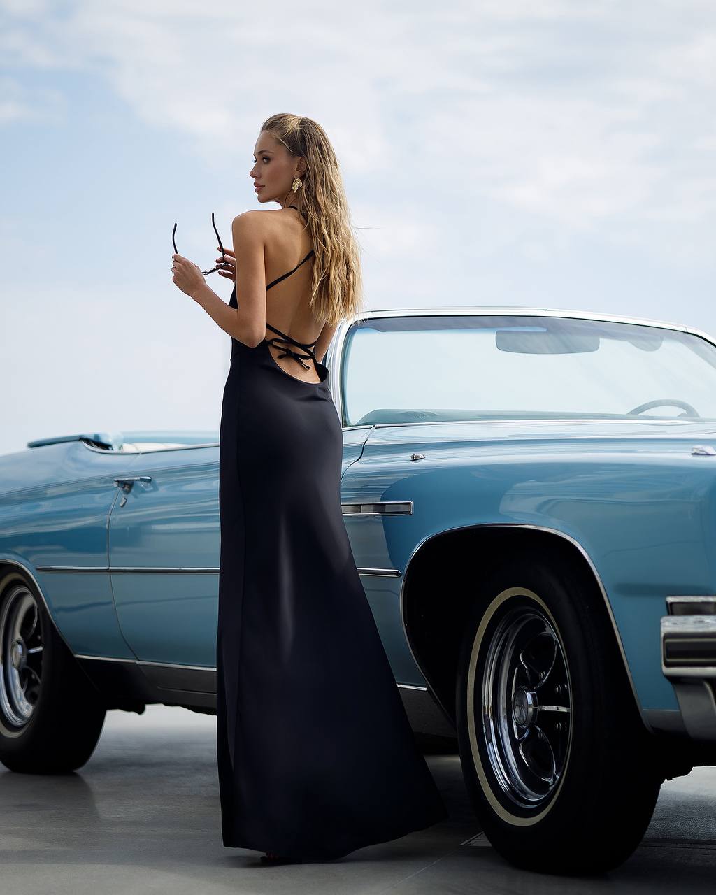 Woman in a black dress standing next to a blue vintage car against a clear sky.