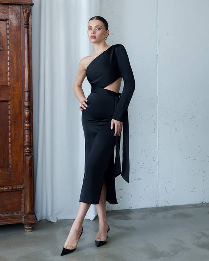 Woman in a black one-shoulder dress standing against a gray wall with a wooden cabinet.