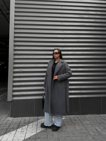 Person wearing a long gray coat standing in front of a corrugated metal wall.