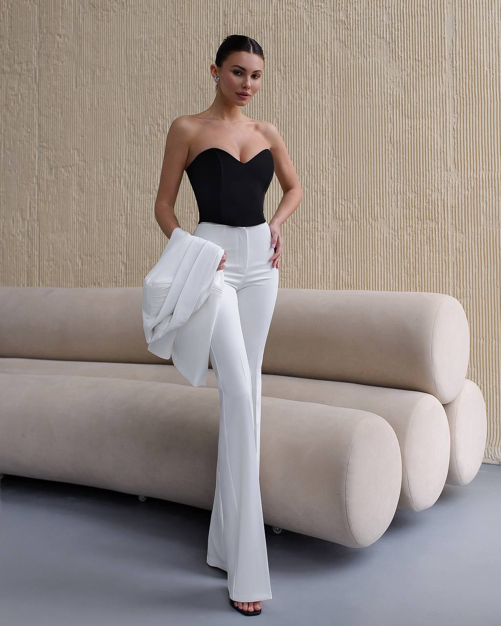 Woman in black top and white pants standing in a modern room with a beige sofa.