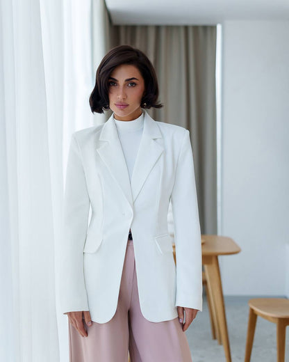Woman wearing a white blazer and light pink pants in a minimalistic room.