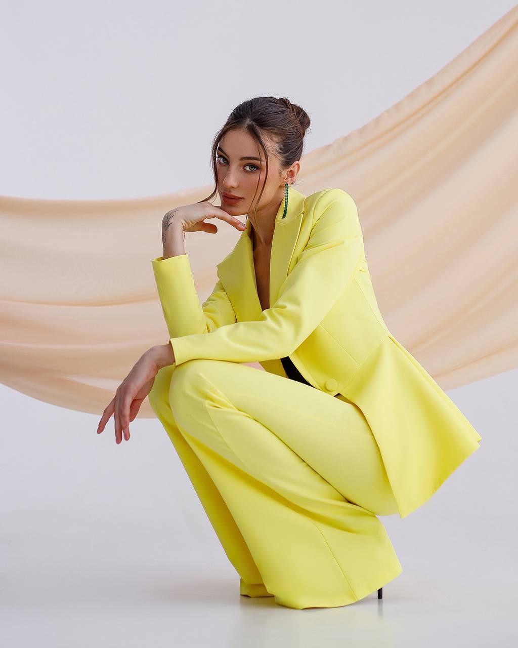 Woman in a bright yellow suit sitting on a white floor with a beige fabric draped behind her.