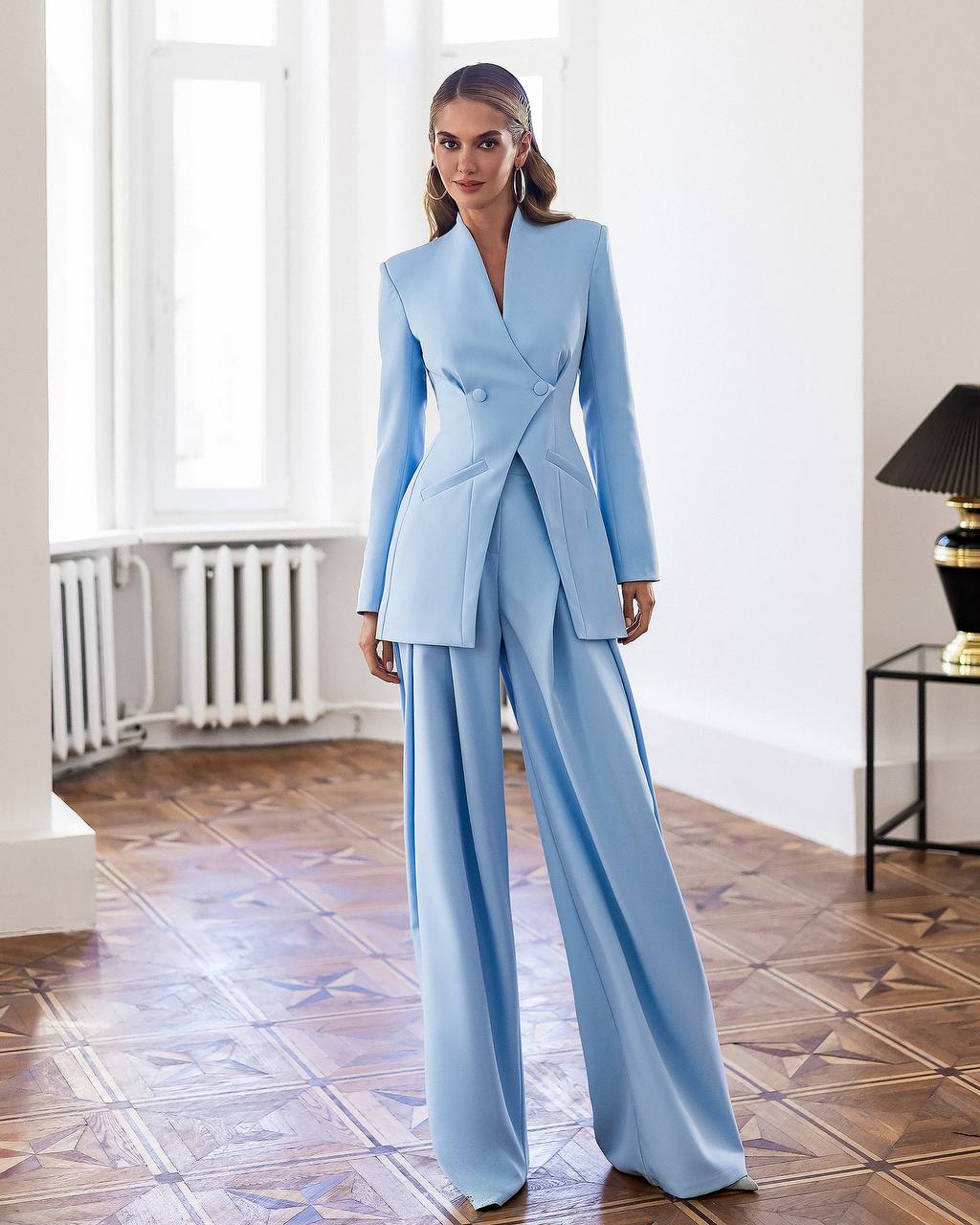 Woman in a light blue suit standing in a room with wooden flooring and white walls.