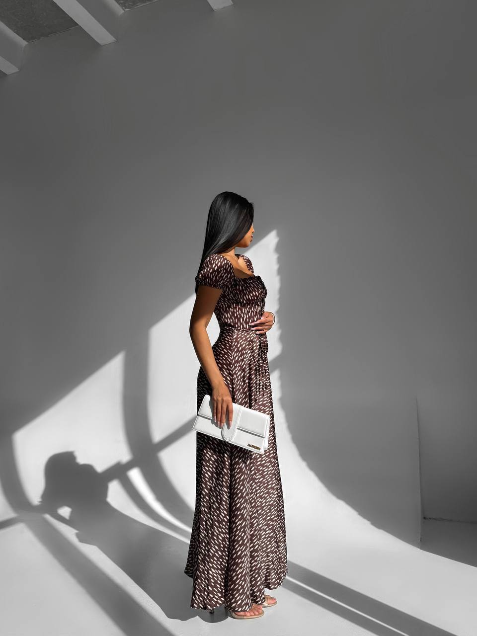 Woman in a long dress holding a clutch in a room with a light source casting shadows.