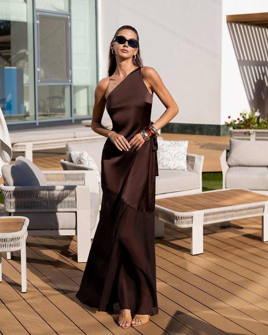 Woman in a brown one-shoulder dress standing on a wooden deck with outdoor furniture.