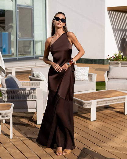 Woman in a brown one-shoulder dress standing on a wooden deck with outdoor furniture.