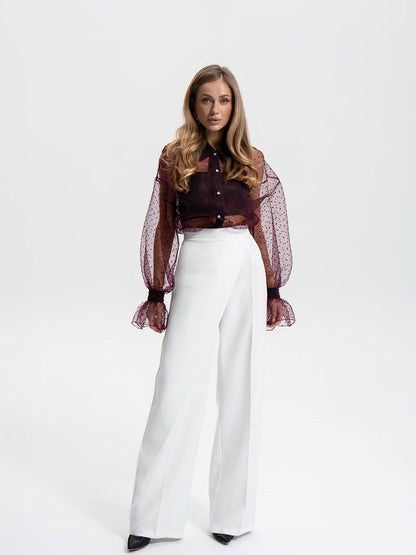 Woman wearing a dark blouse with sheer sleeves and white pants on a white background