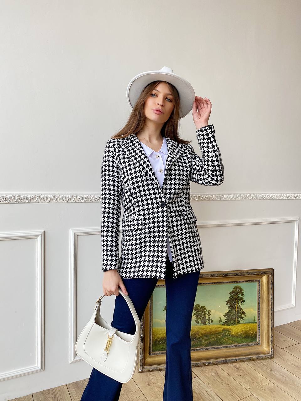 Woman in a checkered blazer and blue pants holding a white handbag, standing in a room with decorative elements.