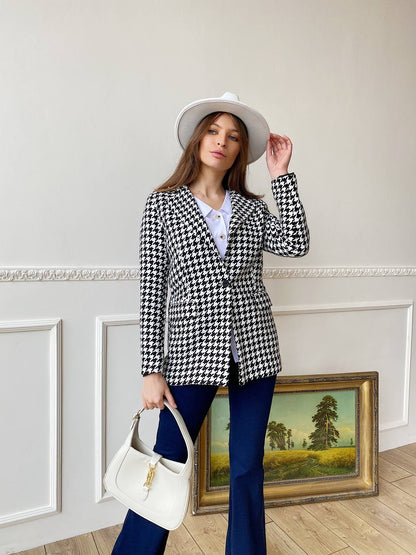 Woman in a checkered blazer and blue pants holding a white handbag, standing in a room with decorative elements.