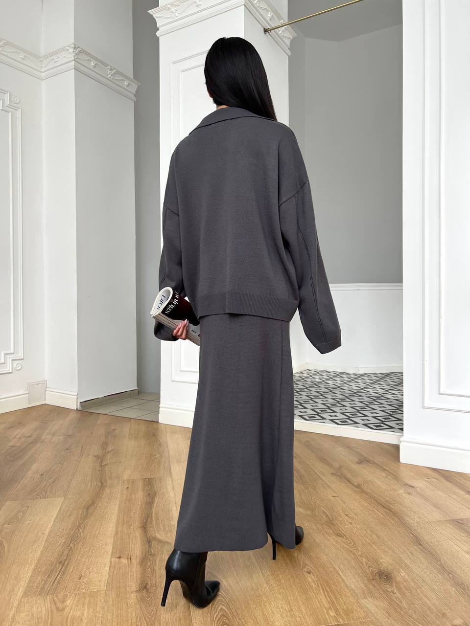 Woman in a gray outfit standing in a room with wooden flooring and white walls.