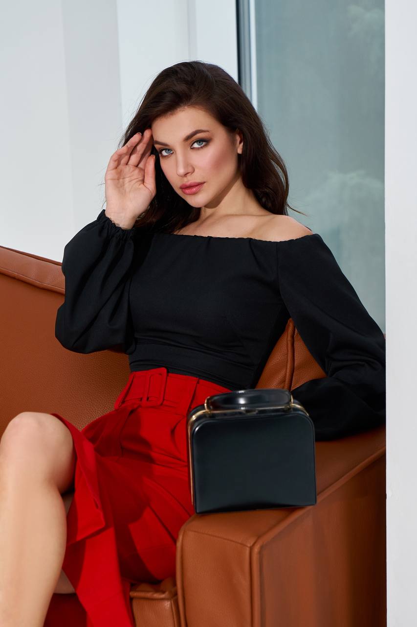 Woman in black top and red skirt sitting on a brown couch with a black handbag.