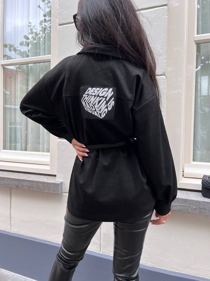 Person wearing a black jacket with text on the back, standing in front of a window.