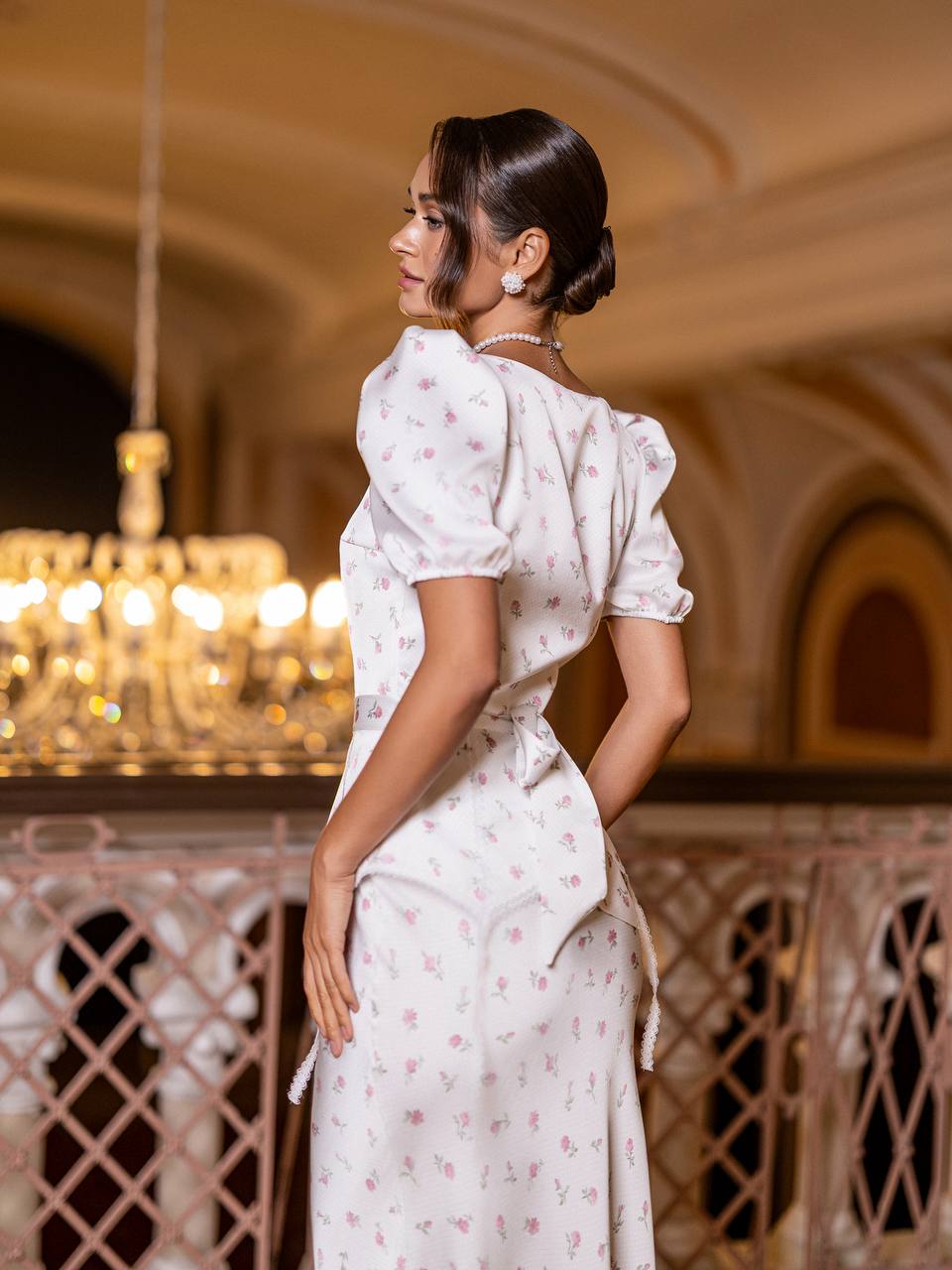 Woman in a white dress with floral patterns standing in an elegant interior setting.